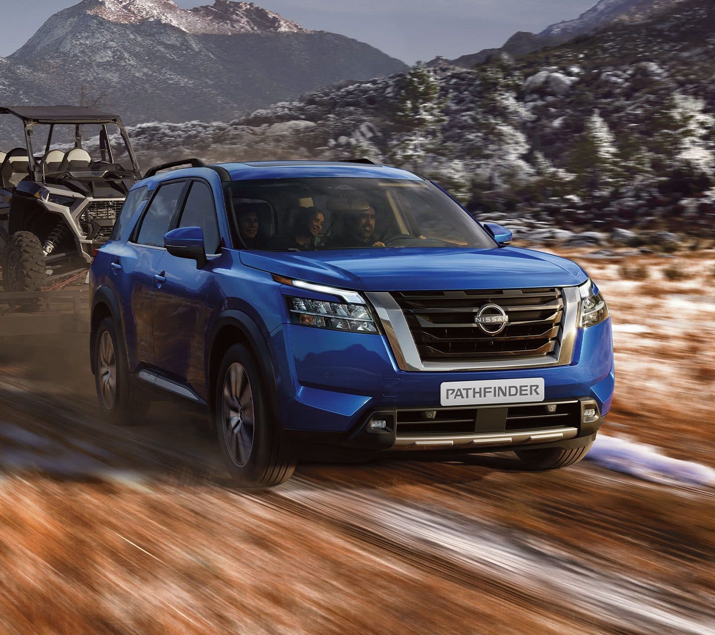 Blue Nissan Pathfinder towing a side-by-side buggy along a dirt track, snow-dusted mountains behind
