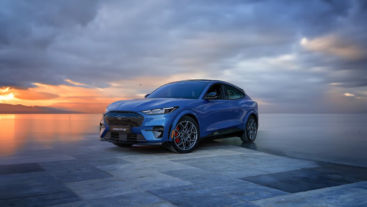 Blue Ford Mustang Mach-E GT parked on a stone waterfront at sunset, front three-quarter view