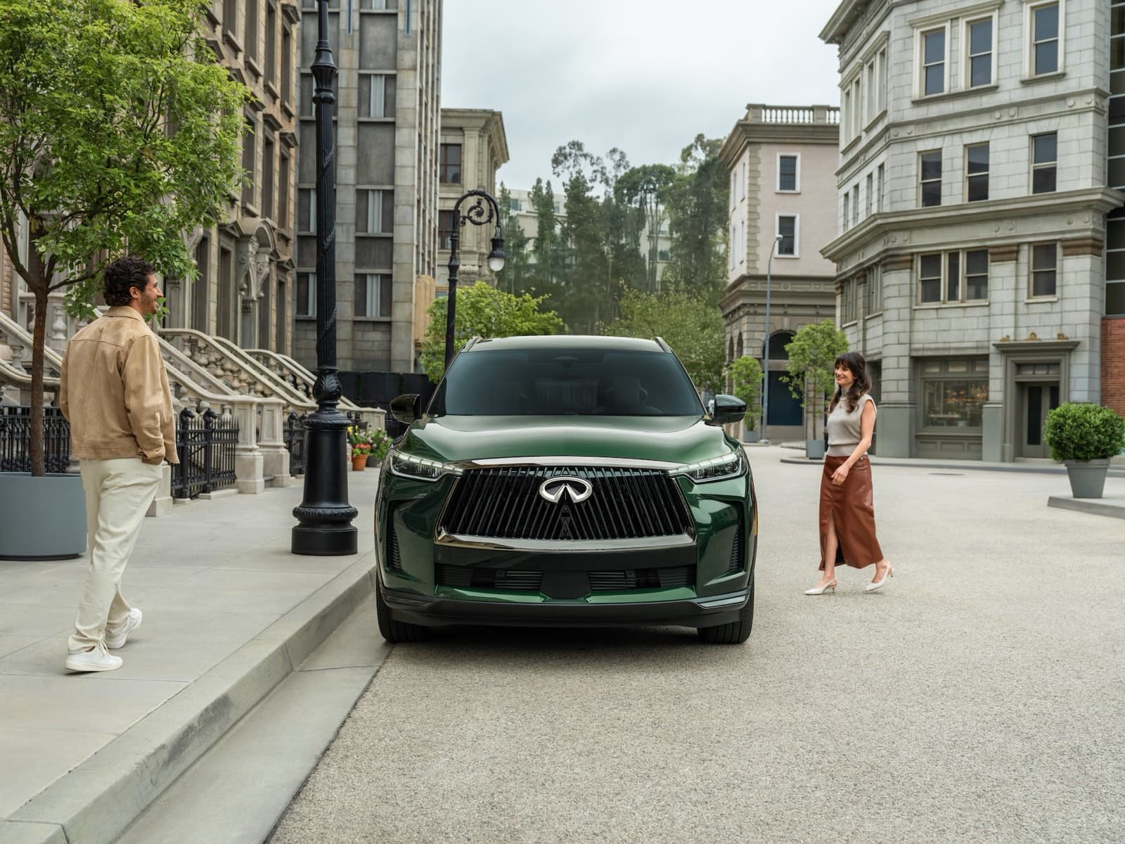 Dark green INFINITI QX60 parked head-on in a city street lined with stone townhouses, a man on the pavement to one side and a woman walking past on the other