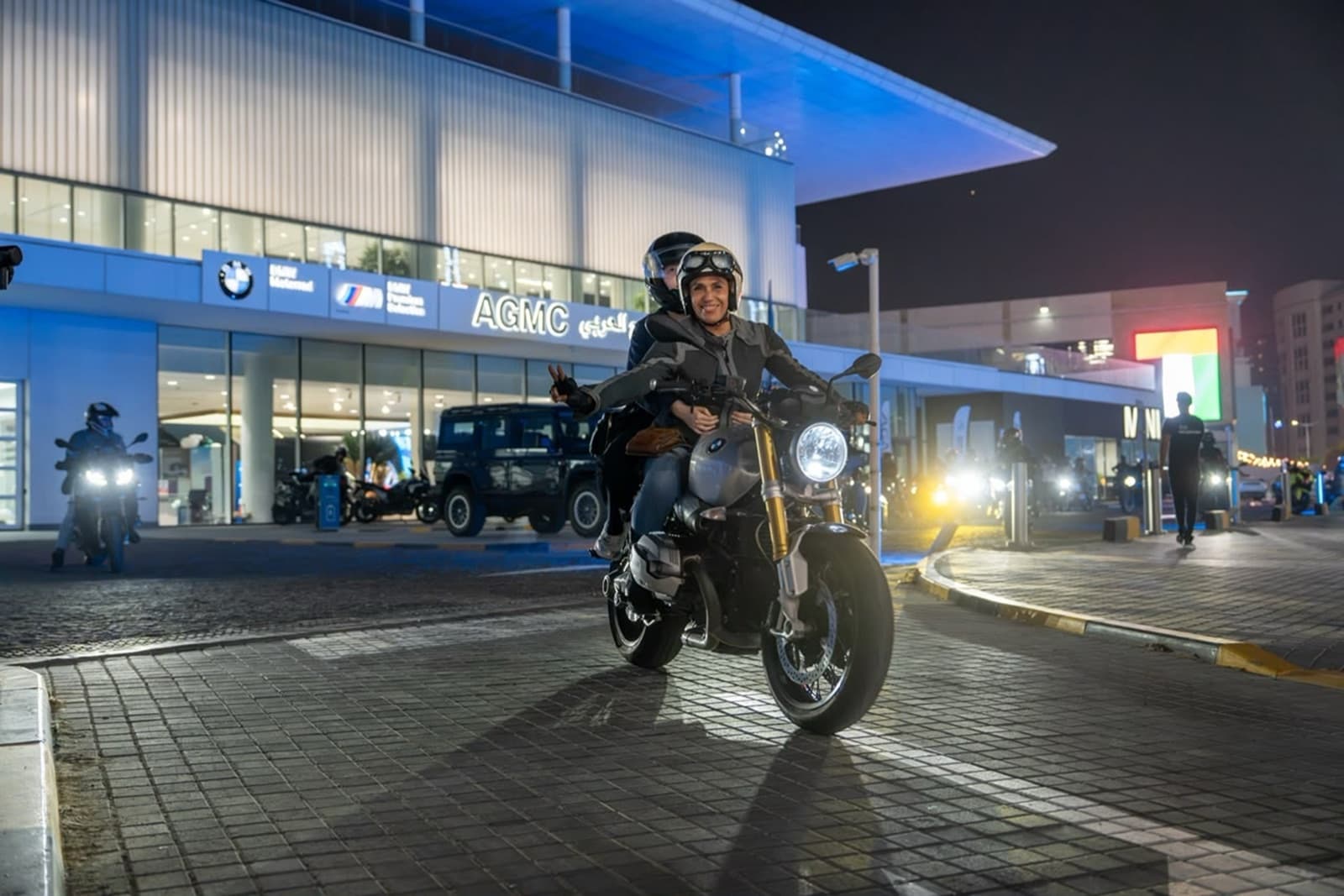 A woman in a helmet and riding jacket rides a BMW roadster away from the lit AGMC Dubai Motor City showroom at night, a pillion passenger behind her and the headlights of other riders following